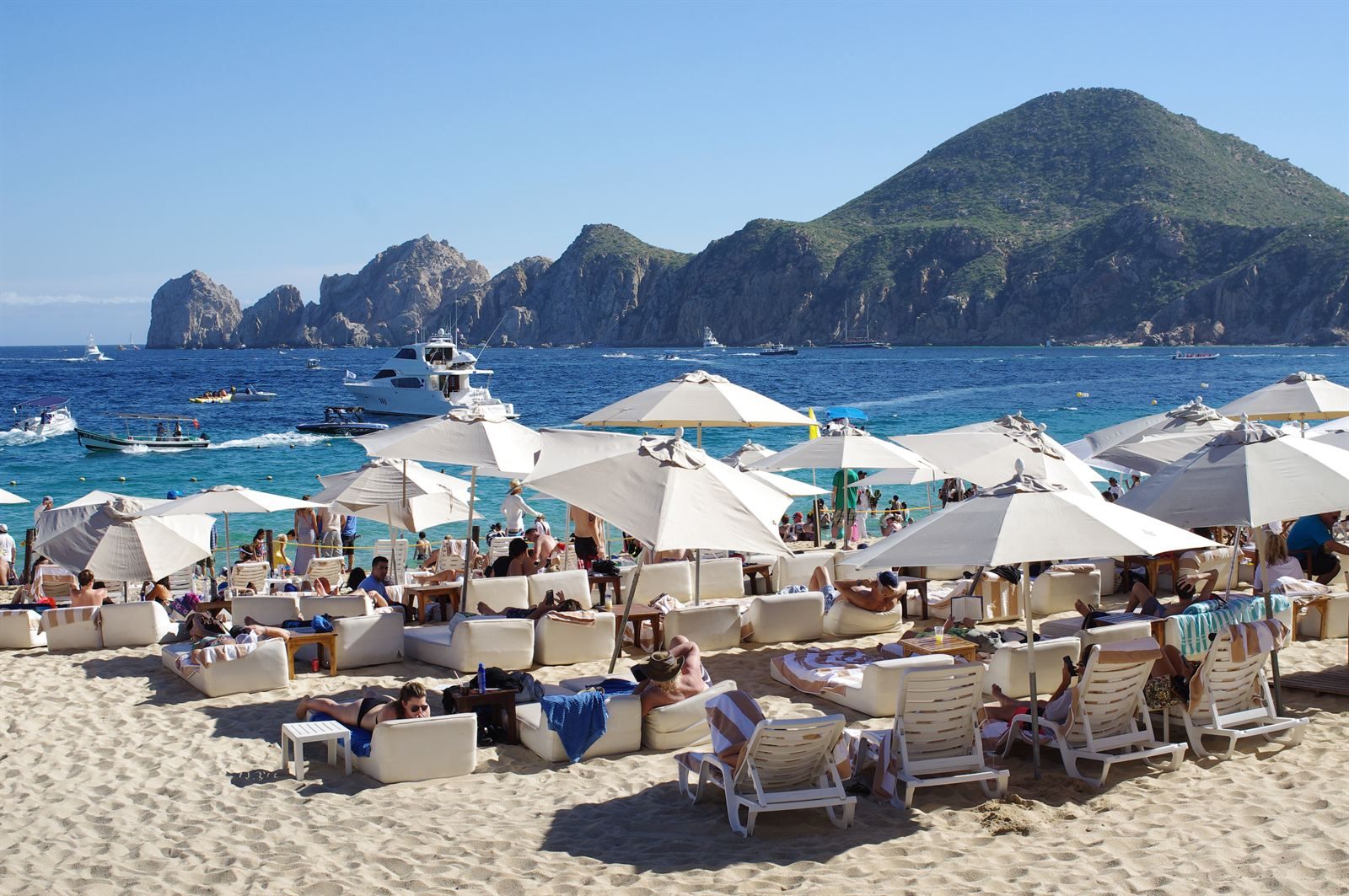 Umbrellas and loungers on a crowded beach in Cabo San Lucas, with boats in the bay and the rocky Land's End headland behind
