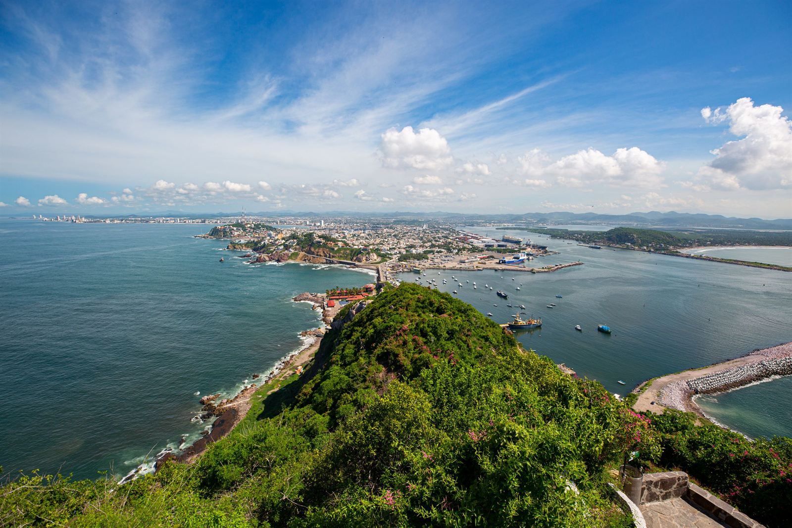 View from a green hilltop over Mazatlan's harbor, breakwaters and the city spread along the coast